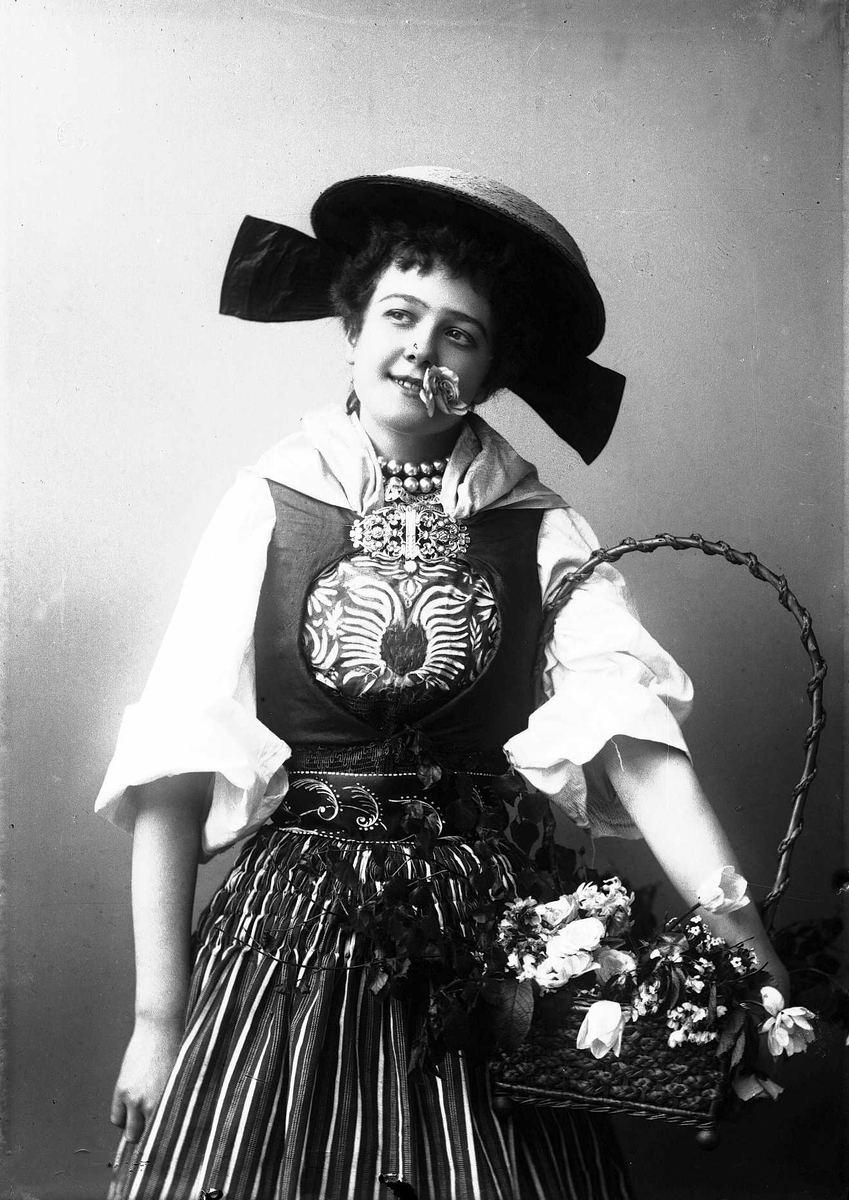 Solveig Lund, Kvinne i drakt og hatt, med blomst i munnen. Holder kurv med blader og blomster under armen.