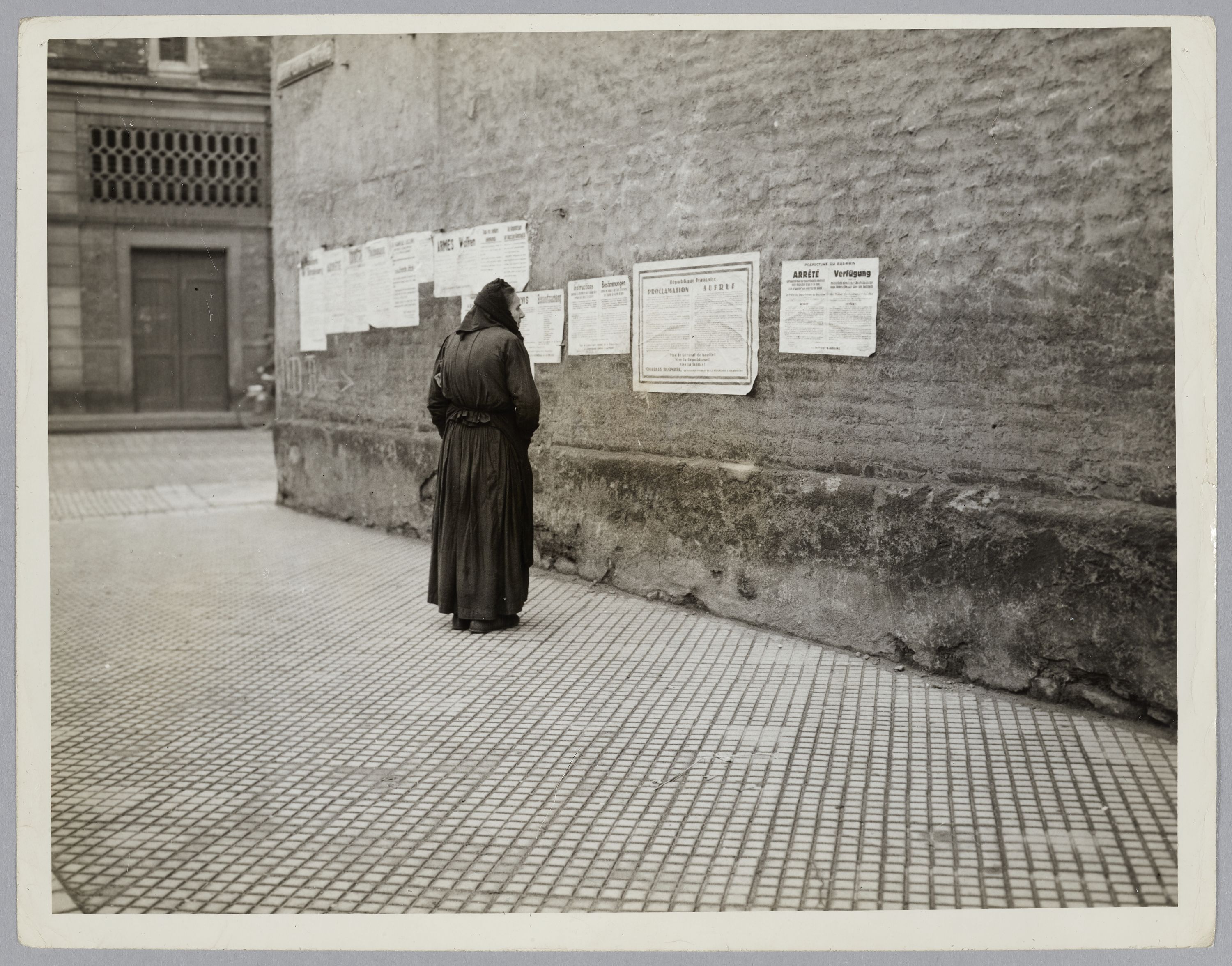 United States Army Signal Corps, Femme âgée en train de lire des affiches bilingues franco-allemandes placardées sur un mur