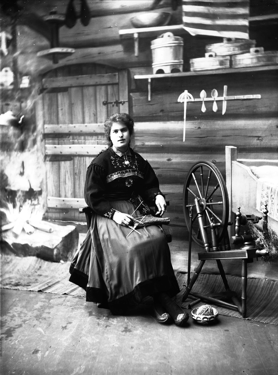 Solveig Lund, Studioportrett av kvinne i nasjonaldrakt. Kvinnen sitter og strikker, ved siden av henne står en rokk. Kvinnen ser på betrakteren. På gulvet står en bolle med et garnnøste. Bakgrunnen er malt og viser seterinteriør. Til venstre står kjelen på ildstedet og til høyre står vegghylle med tiner og en se…