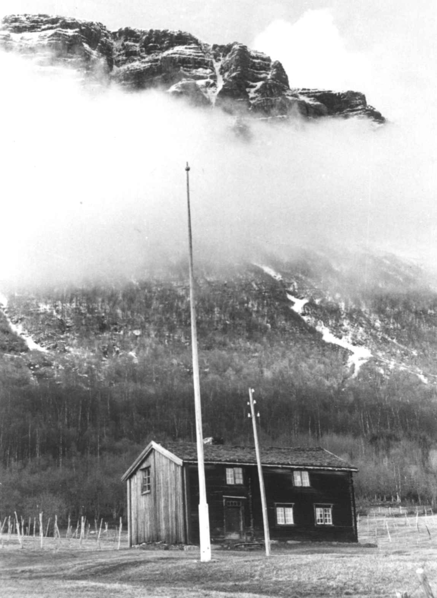 Anna Grostøl, En bygning nedenfor et fjell som stikker opp fra lave tåkeskyer. Rostadal, Målselv 1940-årene.
