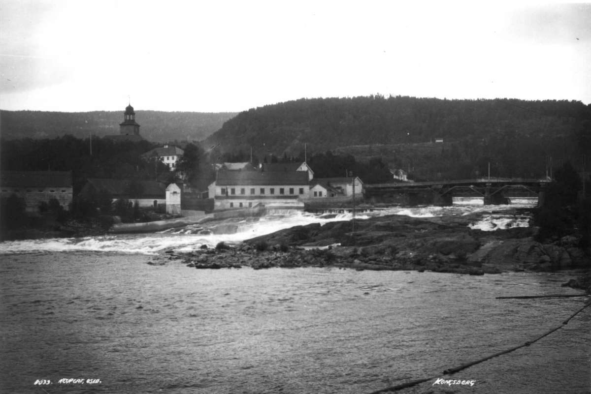 Hermann Christian Neupert, Kongsberg. 1930. Oversiktsbilde.Utsikt over Numedalslågen. Bro. Bebyggelse. I bakgrunnen Kongsberg kirke (1761). Skog rundt byen.