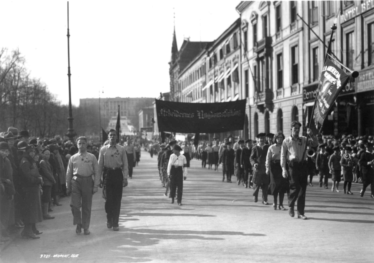 Hermann Christian Neupert, 1. mai-tog, Karl Johans gate. 1930. Slottet i bakgrunnen. Toget på vei mot Stortinget. Menn med faner. Kvinner og barn. Musikkkorps. Tilskuere.