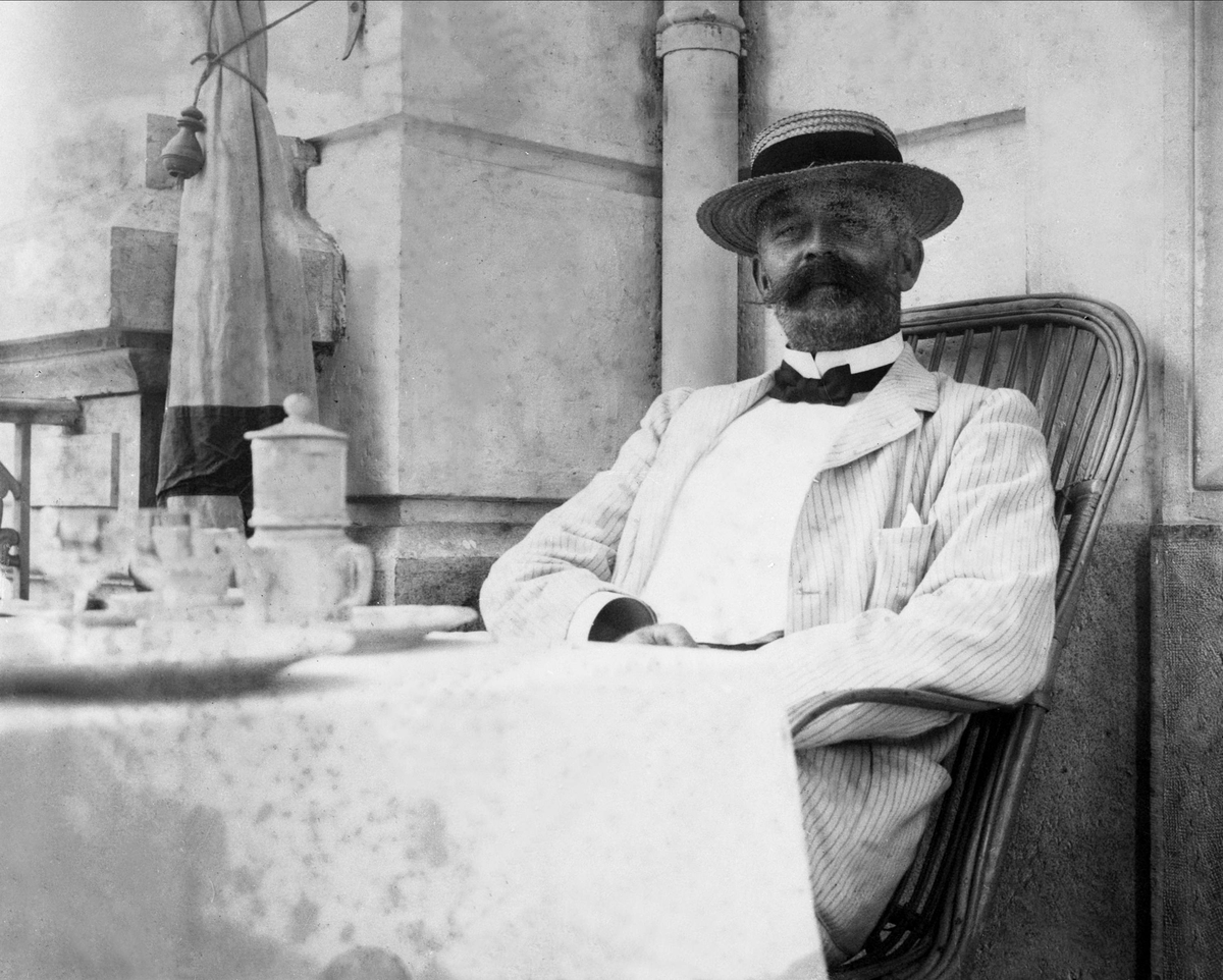 Gustav Borgen, Avfotografering. Mann med stråhatt, sittende på veranda ved dekket bord.