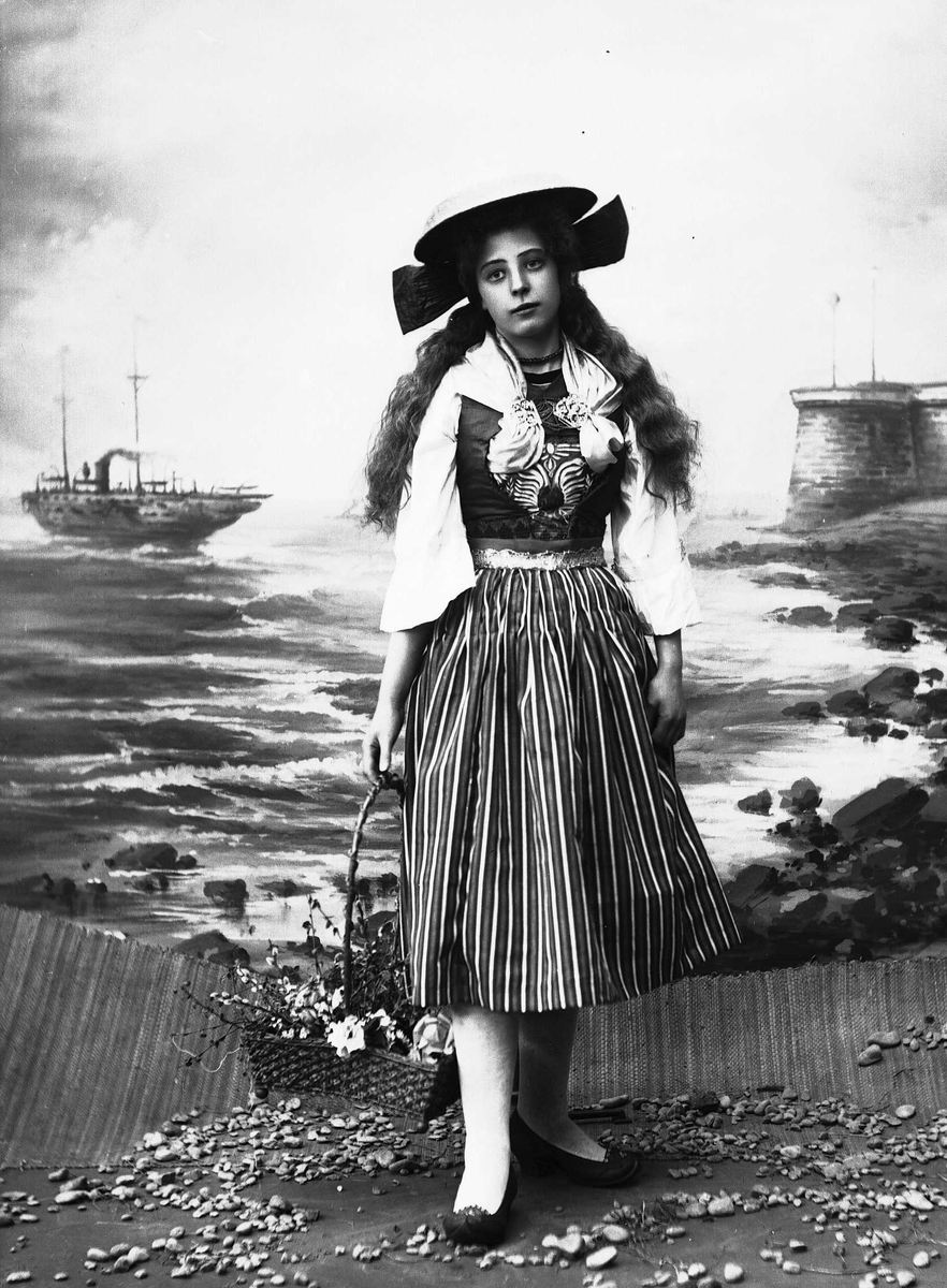 Solveig Lund, Kvinne i drakt og hatt, som holder kurv med grener og blomster. Står foran malt bakteppe med sjø, skip og pir. Steiner på gulvet.
