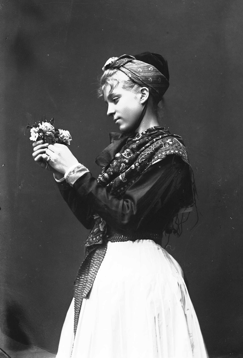 Solveig Lund, 3/4 del studioportrett av kvinne i drakt og oppsatt hår, med sjal rundt skuldrene. Hun holder en bukett blomster i hendene, som hun ser ned på. Nøytral bakgrunn.