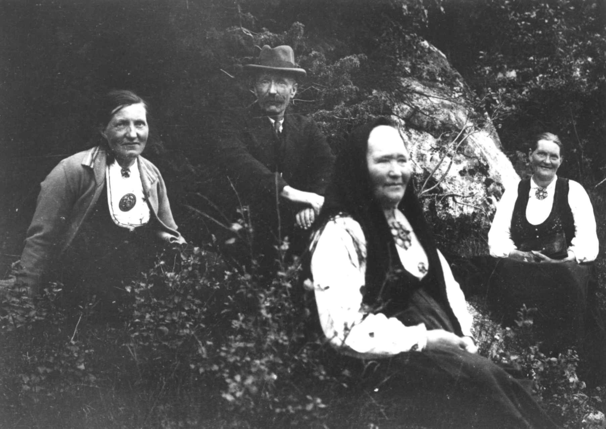 Anna Grostøl, Tre kvinner og en mann, ukjente, i penklær sitter i skogen ved en bergknatt. Ukjent sted 1936.