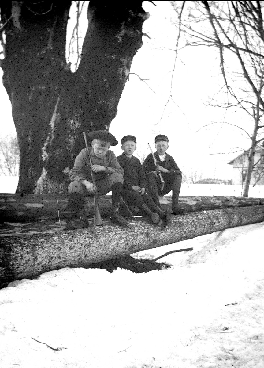 Juliane Fredrikke Langberg, Portrett av 3 gutter med lue sittende på tømmerstokker med geværer i hendene. T.v. Olaf L. Holm, sønn av sokneprest i Hurum, Olaf A. Holm, fotografens svoger. De to andre guttene er sønner av klokker Busland i Hurum. Muligens på Hurum prestegård. Fra malerinne Juliane Fredrikke Langbergs (1856-1930…