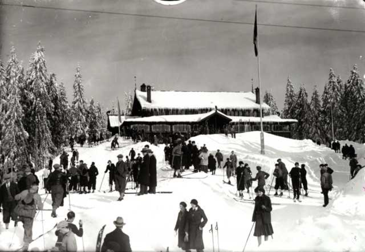 Hermann Christian Neupert, Frognerseteren Restaurant, Nordmarka, Oslo. Vintermotiv. Mennesker på ski foran resturanten. Flaggstang med flagg.