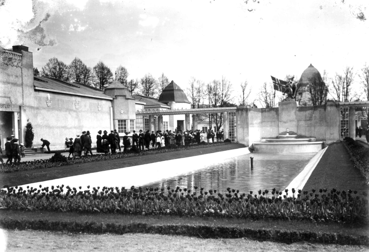 Hermann Christian Neupert, Jubileumsutstillingen på Frogner, Oslo, 1914. Husflidsbygningen til venstre. Basseng. Skulpturen Helhesten av Gunnar Utsond i bakgrunnen. Tulipanbed i forgrunnen. Spaserende menn og kvinner. kvinner. Spaserende menn og kvinner.