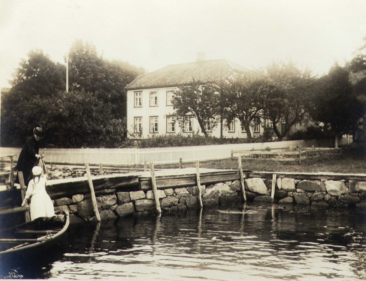 Anders Beer Wilse, Bolighus ved sjøen, Grimstad, Aust-Agder. To kvinner ved robåt i forgrunnen.