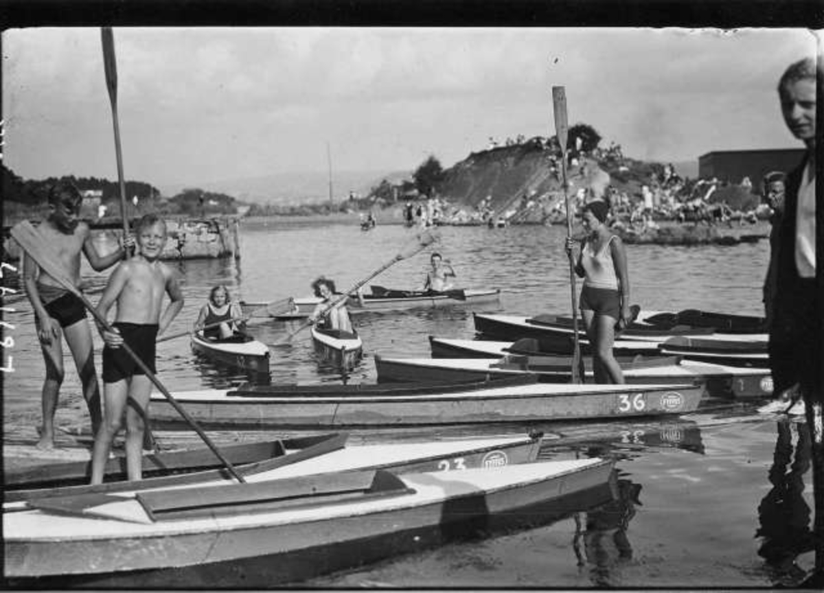 Hermann Christian Neupert, Padling. I Oslofjorden? Menn og kvinner med padleårer. Noen sitter i kajakker. Badende på svabergene i bakgrunnen.