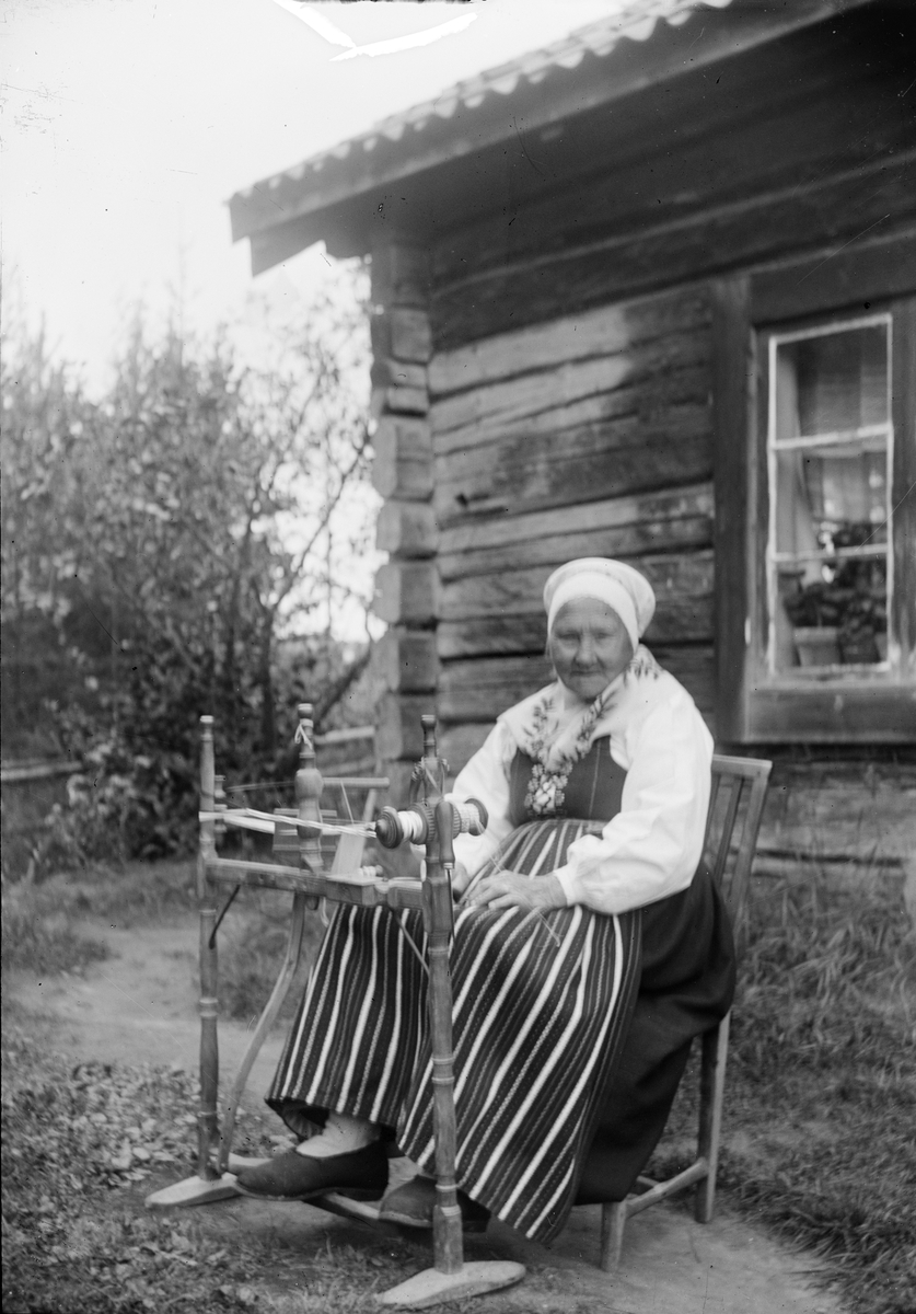 Okänd,, Äldre kvinna, klädd i folkdräkt, ute i trädgård med textilredskap, sannolikt i Dalarna