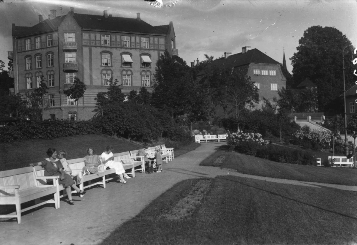 Hermann Christian Neupert, Oslo. Mennesker sitter på benker i en park ved Vestkanttorvet. Bybebyggelse i bakgrunnen. Trær.