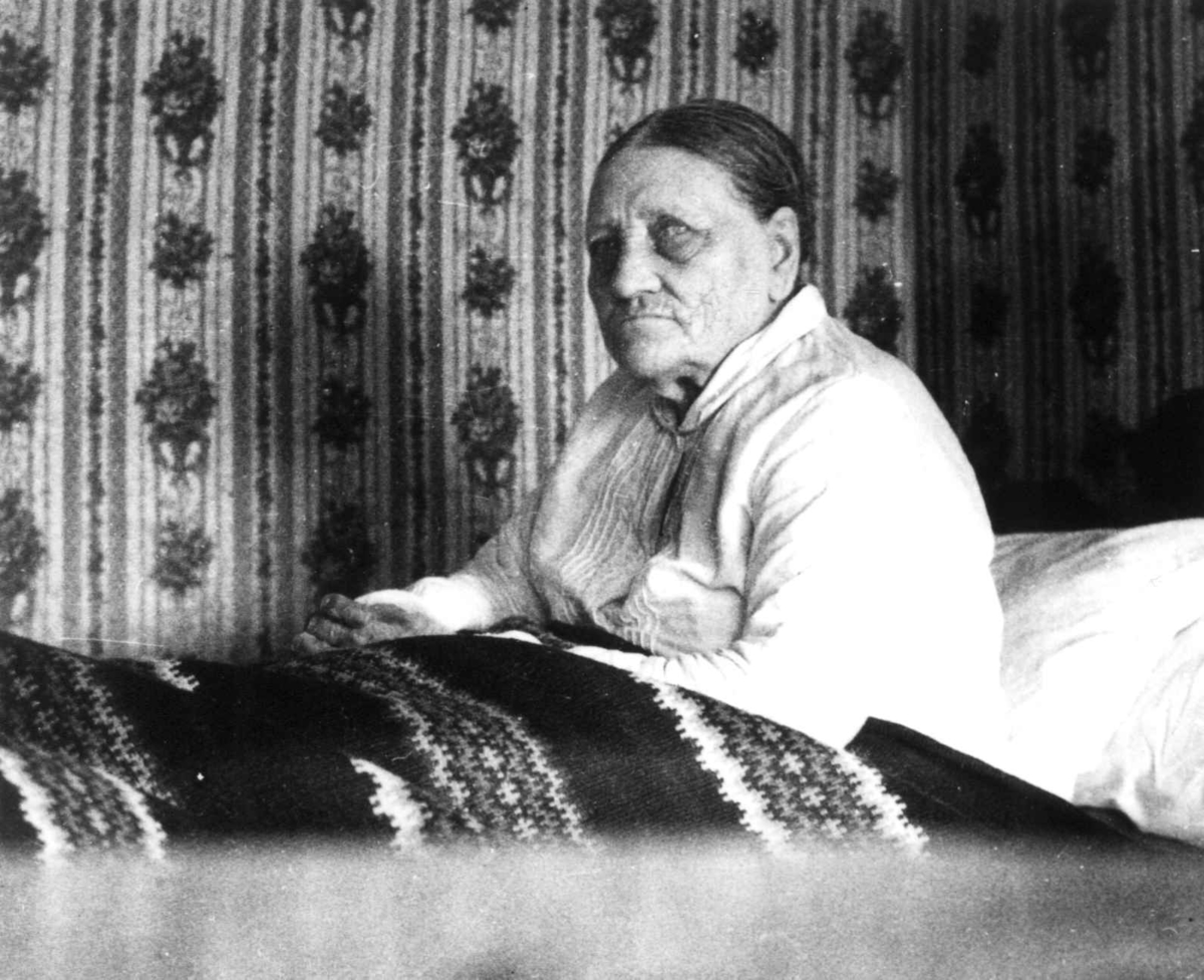 Anna Grostøl, Fru Åselina Eiken sitter i sengen sin. Eiken, Hægesbostad, Vest-Agder 1937.