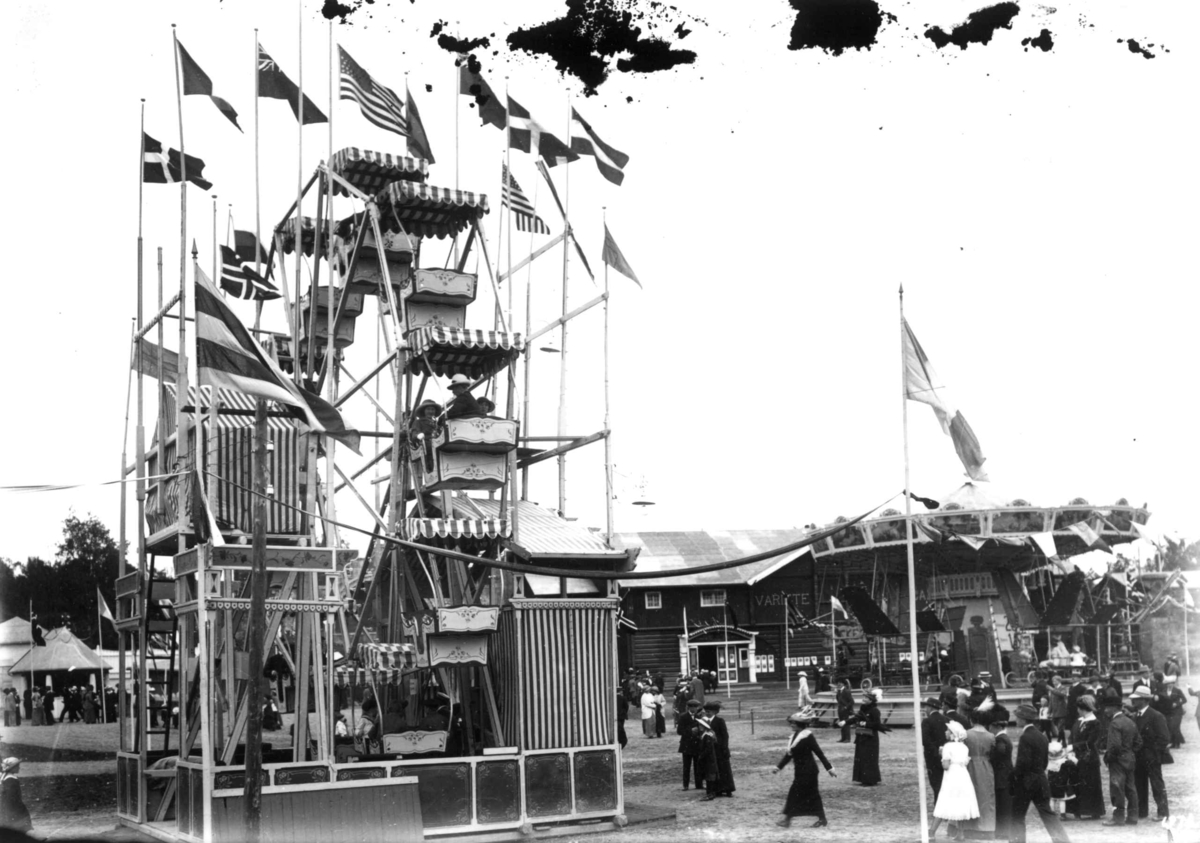 Hermann Christian Neupert, Jubileumsutstillingen på Frogner, Oslo, 1914. Lykkehjul i forgrunnen. I bakgrunnen Tanagra dukketeater. Karusell i forgrunnen. Publikum. Flagg.
