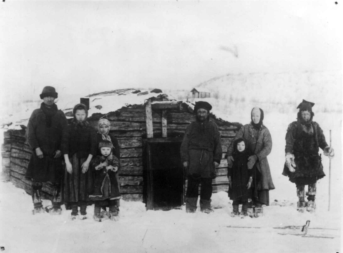 Ellisif Rannveig Wessel, Barn og voksne skoltesamer foran hus, Neiden, Sør-Varanger, Finnmark, ca. 1904. Personer oppgitt: Ondre Jakovitsj Romman (1858-1926), Nikolai Ondrevitsj (1839-1920) og Ivan Ondrevitsj.
