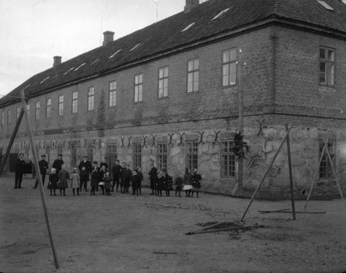 Hermann Christian Neupert, Postkort. Artillerikasernen fra 1733, Fredrikstad festning. Granittmur i 1. etasje og spekket teglstein i overetasjen. Voksne og barn på plassen foran bygningen. Fot. ant. Neupert 1930.