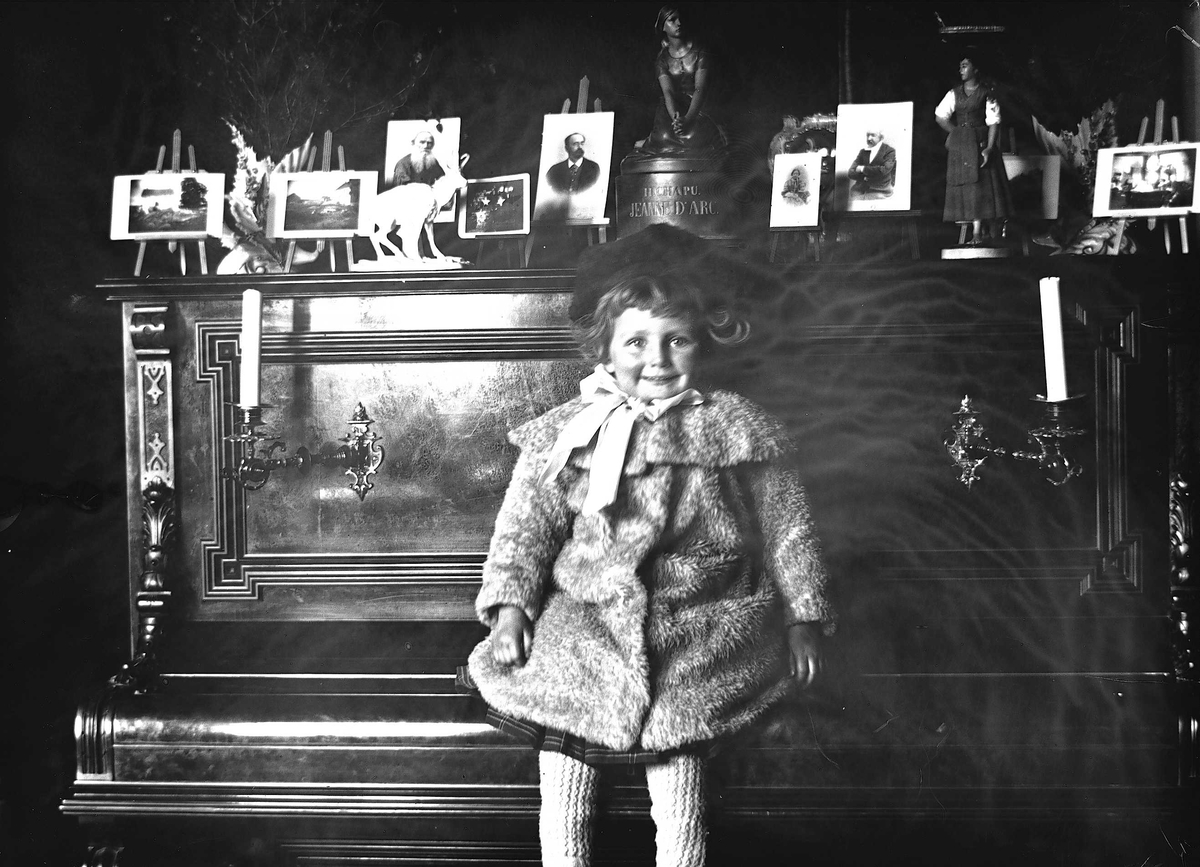 Axel Quinsgaard Wiborg, Gudrun Q. Wiborg sitter på pianolokk i Meltzers gate 9, Oslo, 1899. Fotografier i ramme plassert på pianoet.