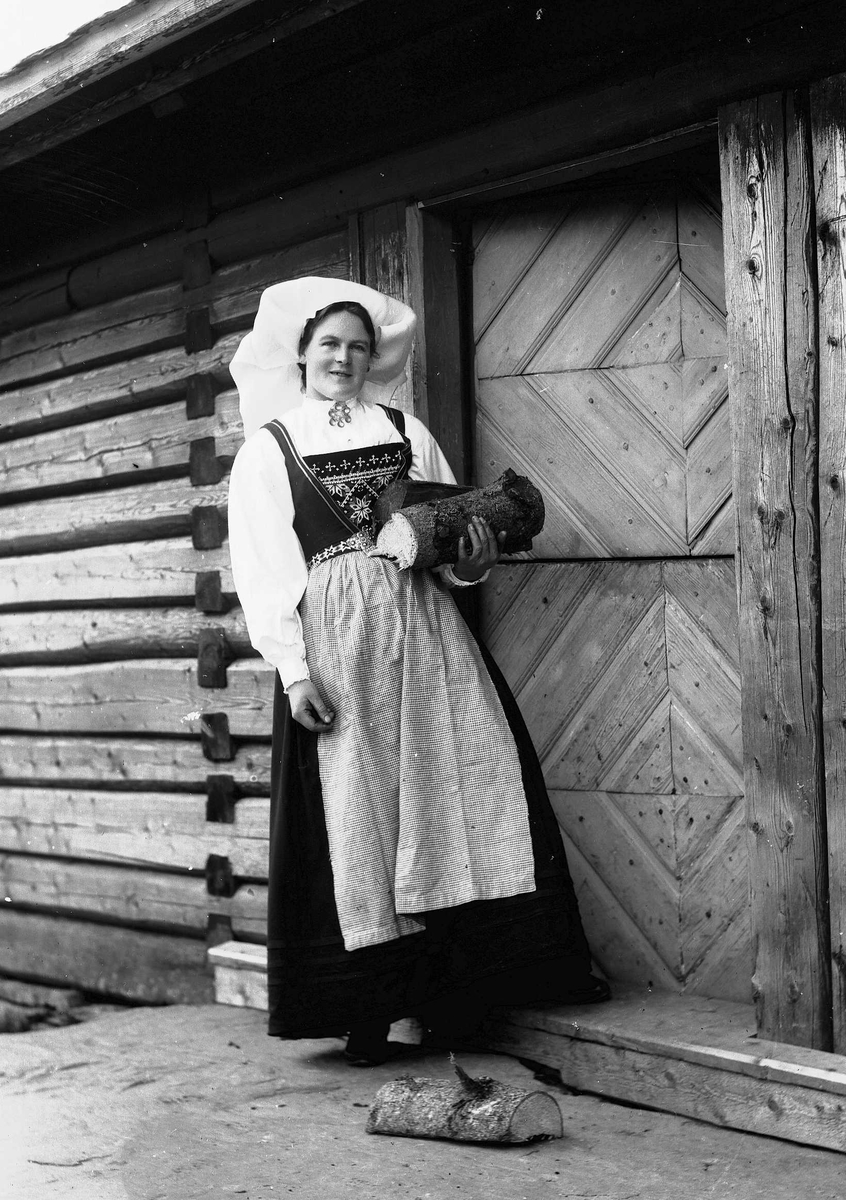Solveig Lund, Kvinne i Hardangerdrakt står i inngangsdøren til seter med vedkubber i armen.
