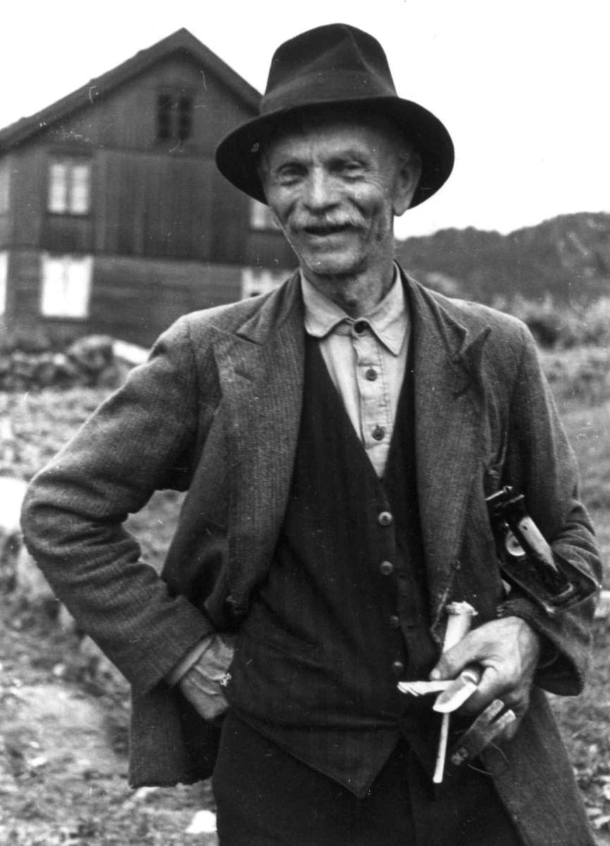 Anna Grostøl, Portrett av Sigmund Rudlende med verktøy i den ene hånden. En bolig i bakgrunnen. Eiken 1941.