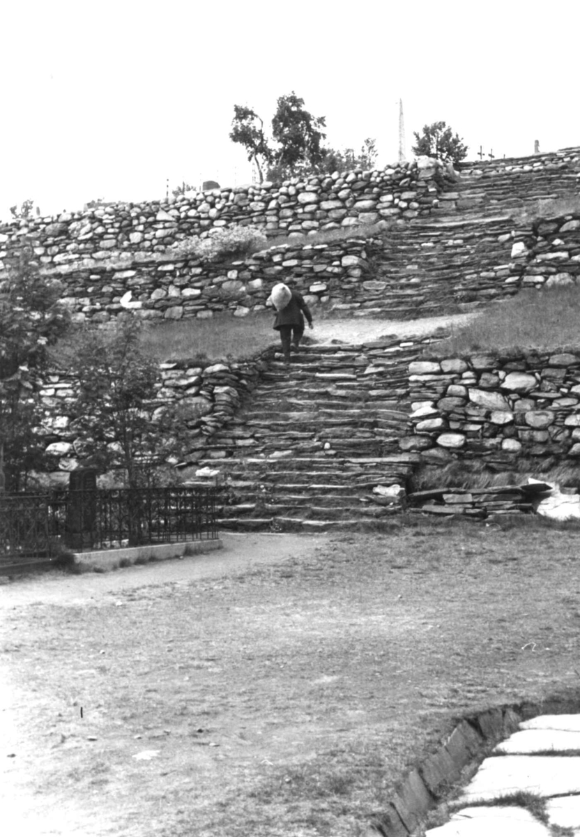 Anna Grostøl, En mann med sekk går oppover en steintrapp bygd i etasjer, antagelig på en kirkegård. Røros 1938.