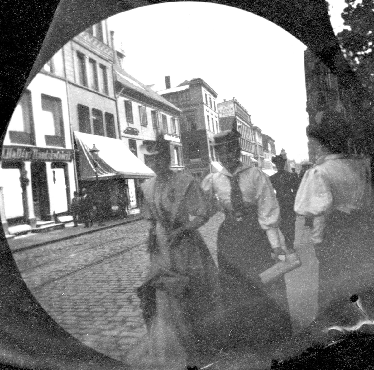 Fredrik Carl Mülertz Størmer, Frk, Ringnes og Mona Jensen spaserer i bygate, Oslo. Brostein, bygårder og butikker.