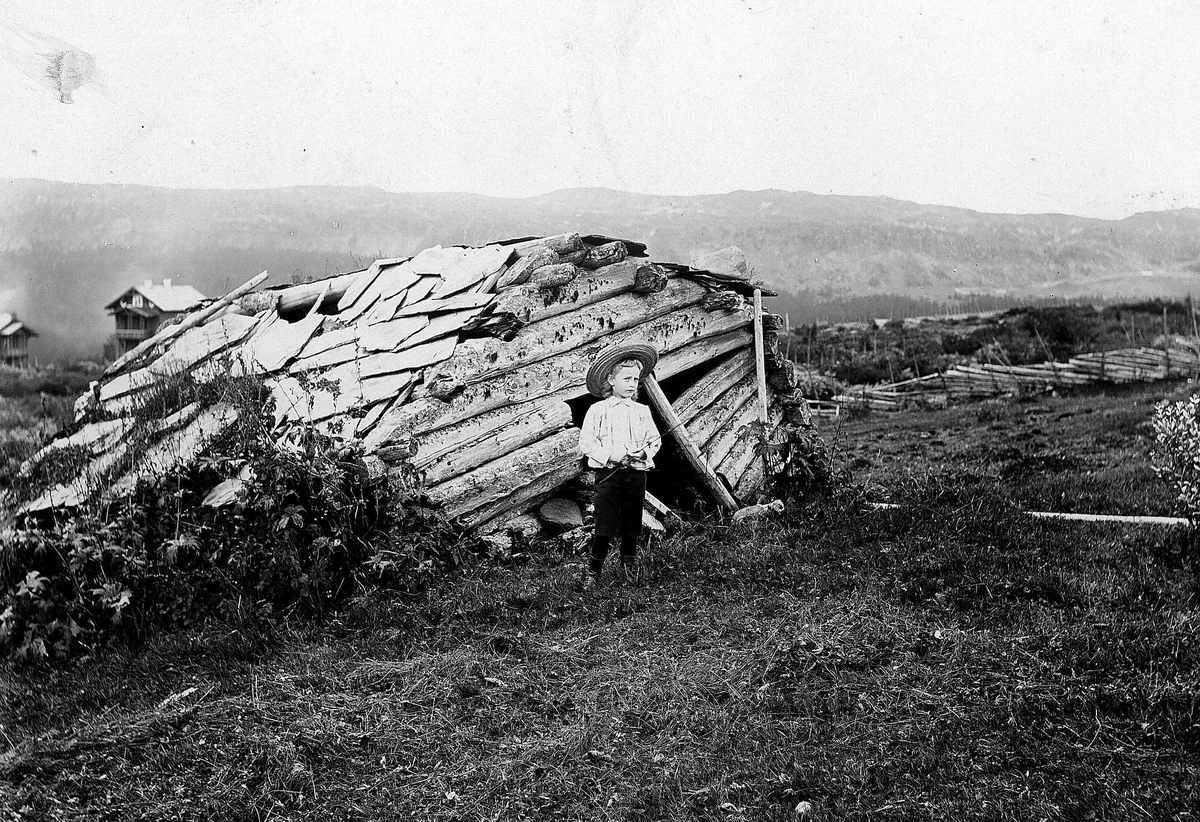 Robert Collett, Gutt med vidbremmet hatt og hvit skjorte foran sammenfalt uthus med skifertak, ukjent sted. Muligens seterområde i fjellterreng. Serie tatt av Robert Collett (1842-1913), amatørfotograf og professor i zoologi.