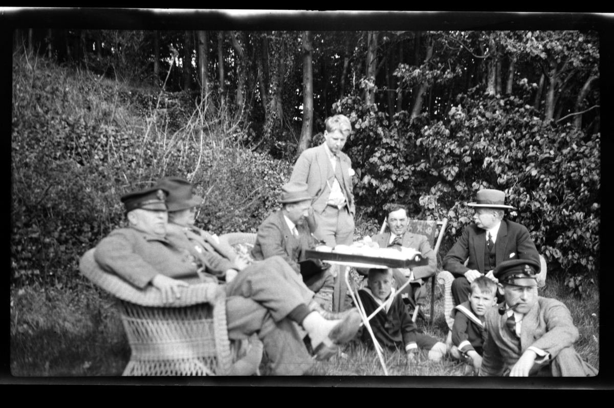 Rolf Sr. Sundt, Gruppe av unge og gamle menn samlet rundt et bord i hage. Fra venstre Halfdan, Trygve, Finn og Bjarne Sundt, Johan Buratti, Christen og Bjørn Reymert og Peter Sundt. Ukjent gutt under bordet. Fotografert 1928.