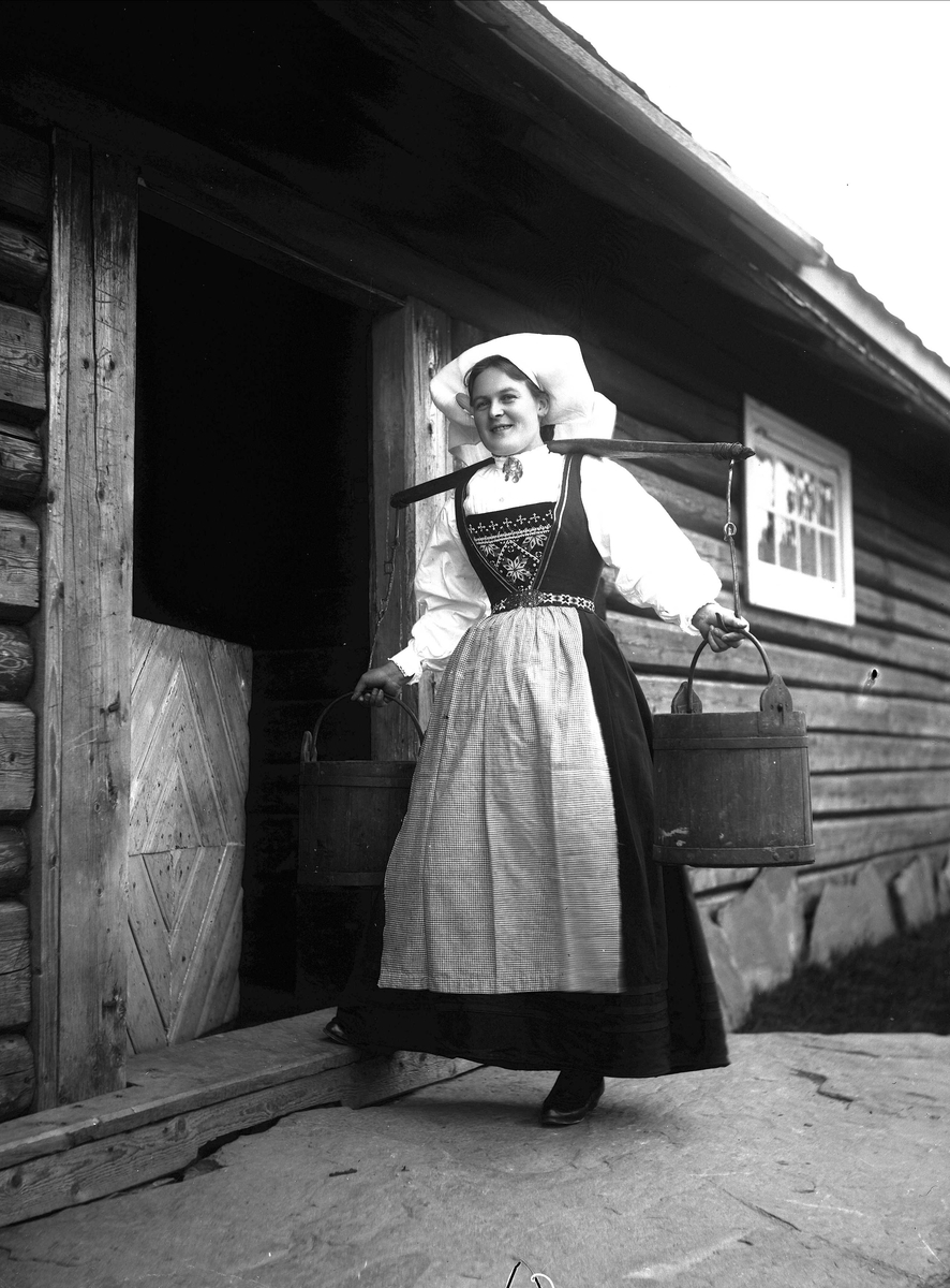 Solveig Lund, Kvinne i Hardangerdrakt står foran dør til seter. Bærer på to vannbøtter.