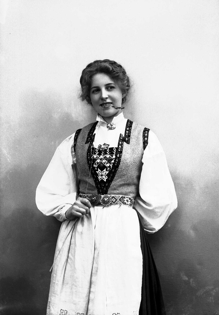 Solveig Lund, 3/4 dels studioportrett av kvinne i drakt, med oppsatt hår. Hun biter på et strå hun holder i høyre hånd. Hun er avfotografert mot en nøytral bakgrunn.