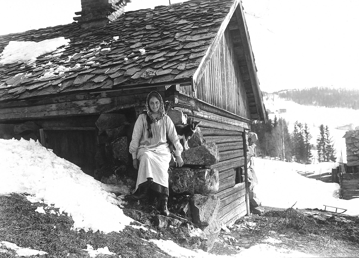 Robert Collett, Pike/kvinne (budeie?) med geit sittende utenfor uthus, antatt fjøs vinterstid, ukjent sted. Huset har skifertak. Jenta er kledd i skaut, forklekjole og snørestøvler. Serie tatt av Robert Collett (1842-1913), amatørfotograf og professor i zoologi.