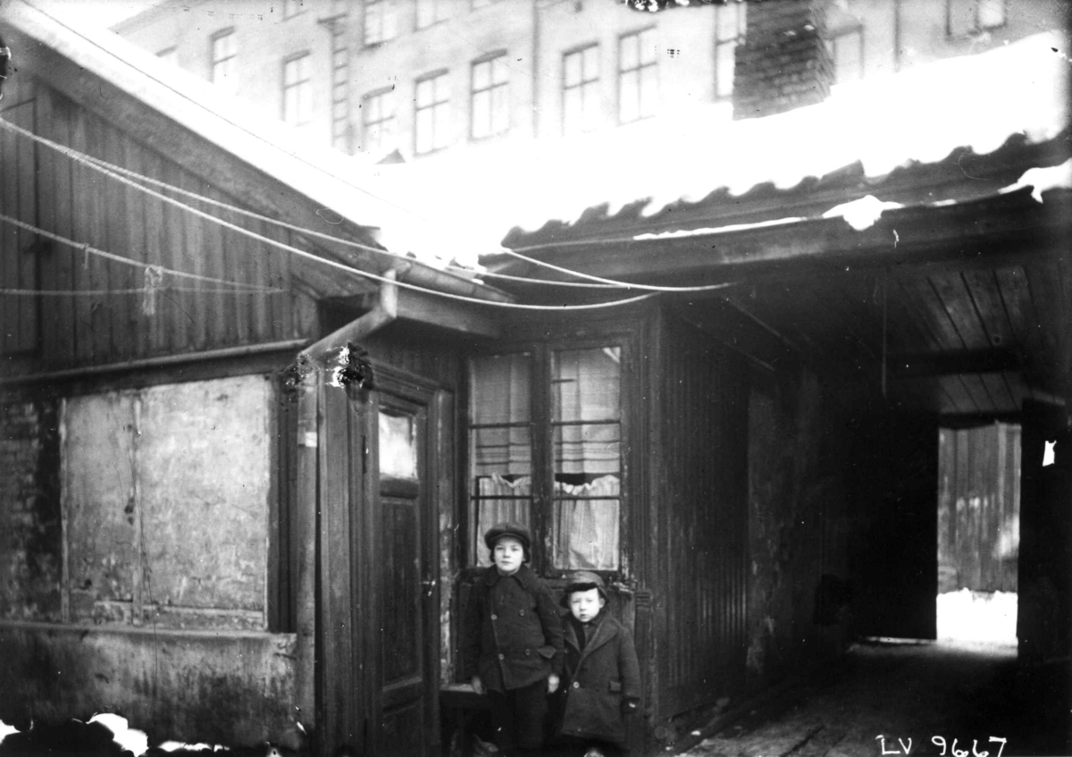 Nanna Broch, Gårdsrom, bolighus, lav trebebyggelse med høye murgårder i bakgrunnen. Barn poserer foran inngangsdør. Fra boliginspektør Nanna Brochs boligundersøkelser i Oslo 1920-årene.