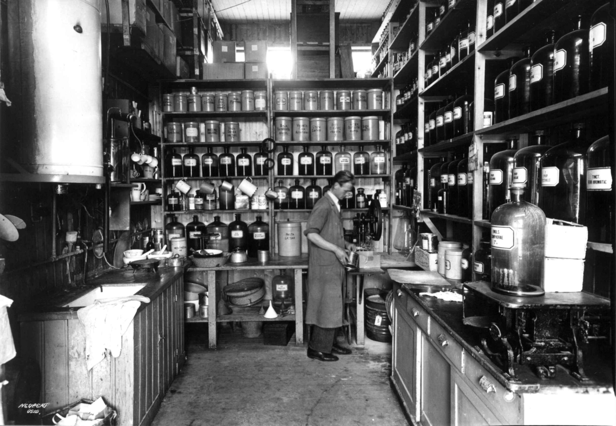 Hermann Christian Neupert, Rådhusapoteket, Oslo. 1935-1937. Provisorisk brakke. Laboratoriet. En mann i arbeid. Hyller med flasker og krukker med ingredienser.