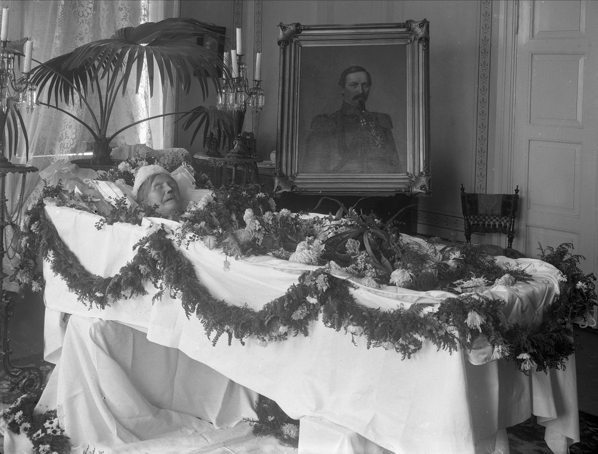 Gustav Borgen, Post mortem-fotografi. Kvinne i pyntet kiste, maleri av avdød ektemann i uniform i bakgrunnen.