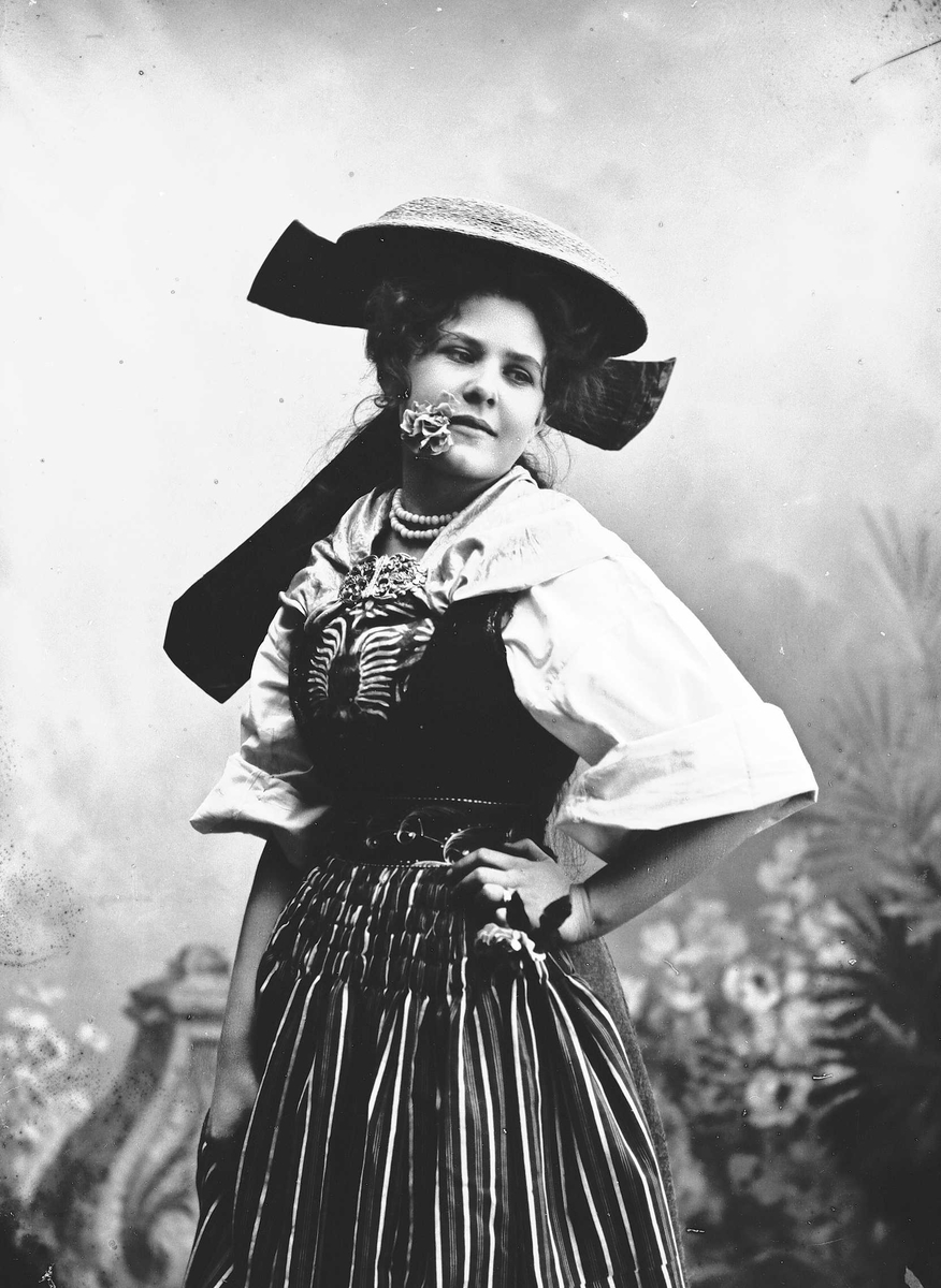 Solveig Lund, Kvinne i drakt og hatt, med blomst i munnen, plassert foran bakteppe med bregner og blomster.