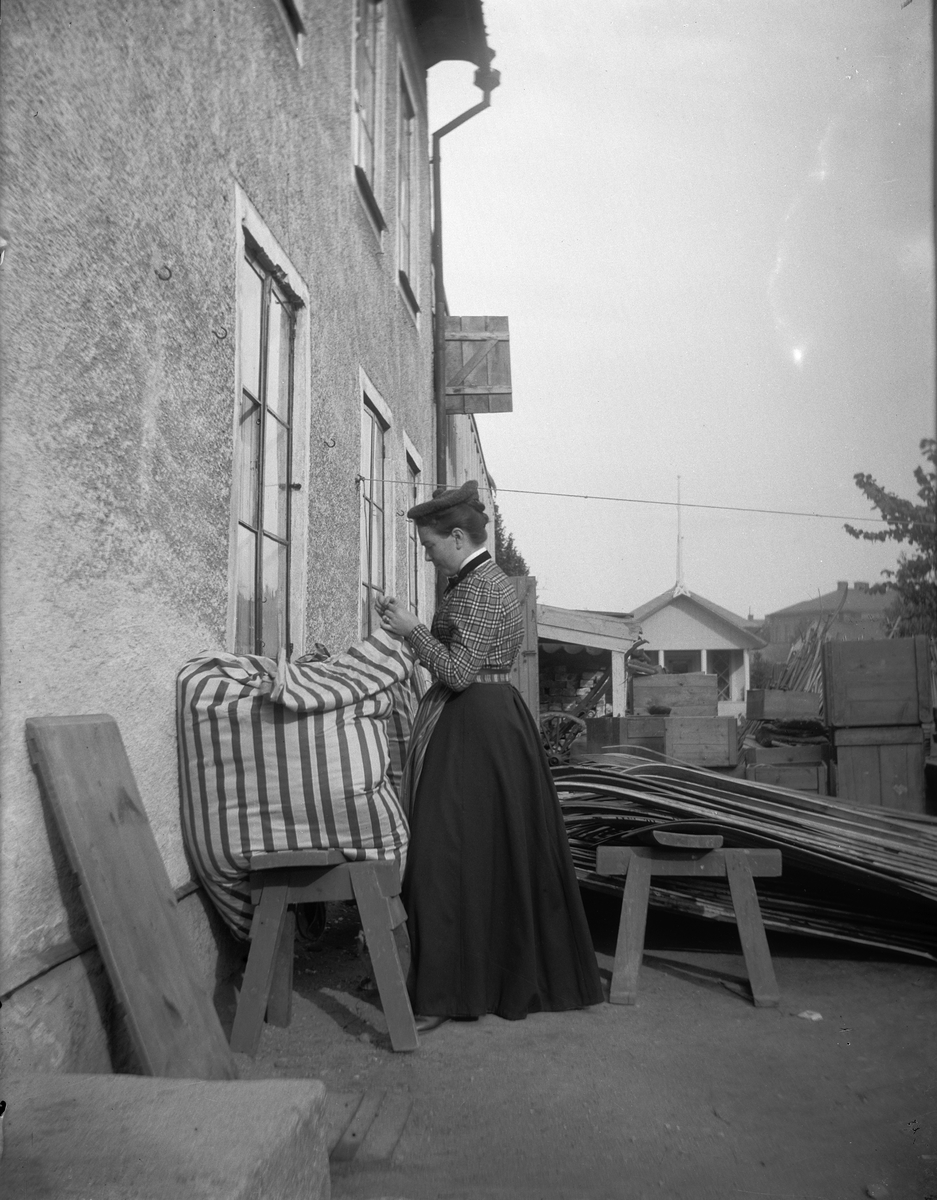 Carl Liljefors, Elin Liljefors står utanför makens kruthandel och syr på ett bolster, Svartbäcksgatan 37, Svartbäcken, Uppsala 1901