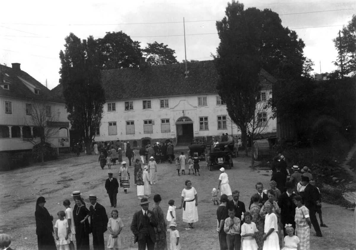 Hermann Christian Neupert, Son, Vestby, Akershus 1924. Festkledde mennesker foran hvite bygninger. Spinnerigården i bakgrunnen. Brant 17. mai 1976. Biler. Trær.