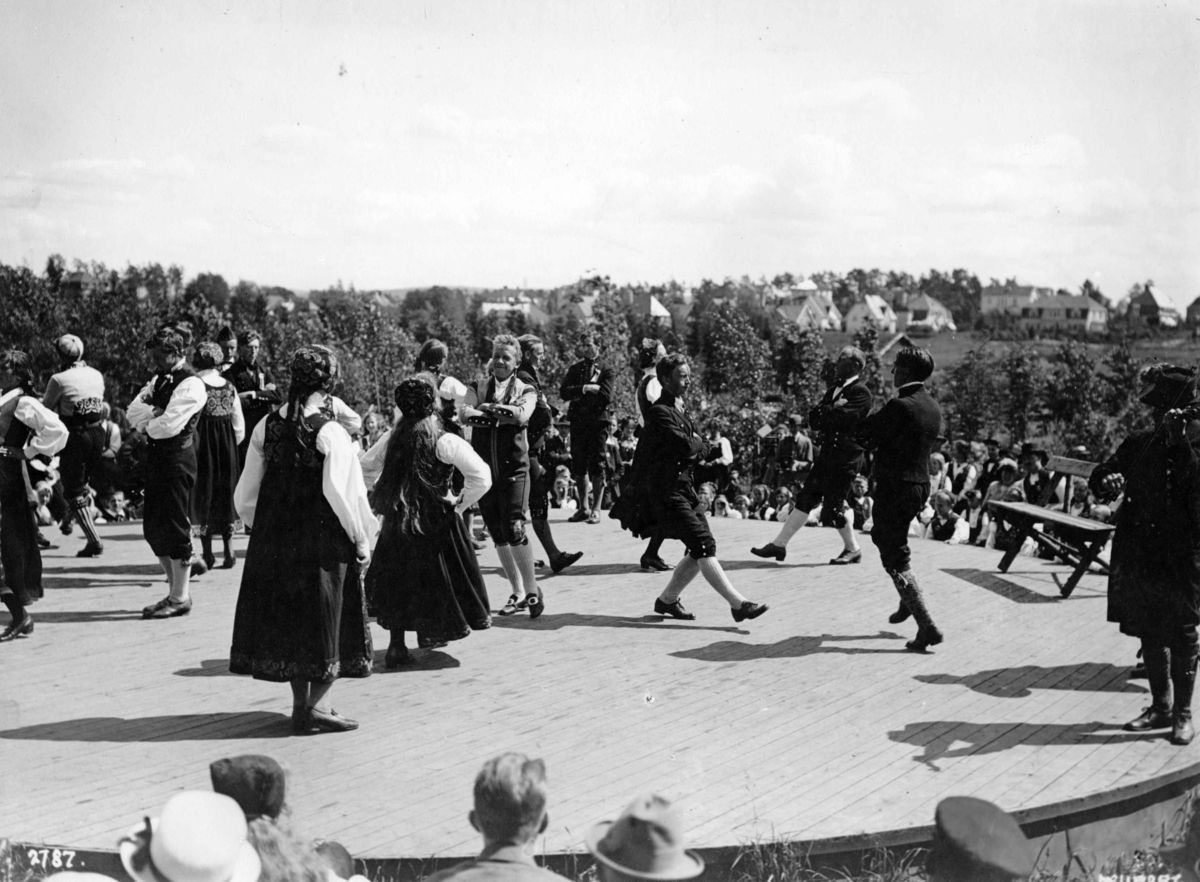 Hermann Christian Neupert, Folkedansoppvisning på en scene. Publikum. Trær og bebyggelse i bakgrunnen.