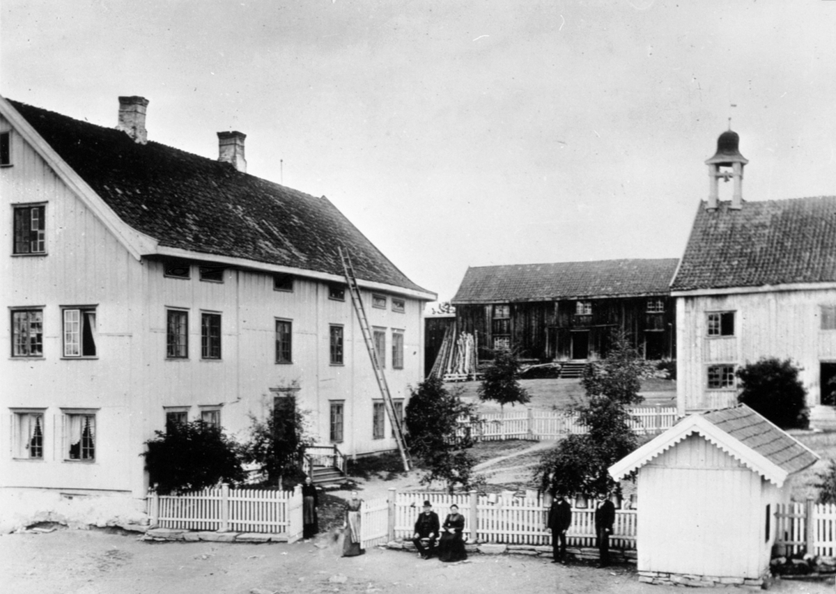 Anno Domkirkeodden, Eksteriør, gardsbruk. Hjelmstad gård, Ringsaker. Hovedbygning, militærkaserne til offiserer, drengestue med klokketårn, brønnhus. Marie f.1834 og Johannes Hjelmstad f.1832 sitter på krakken, resten er ukjente.