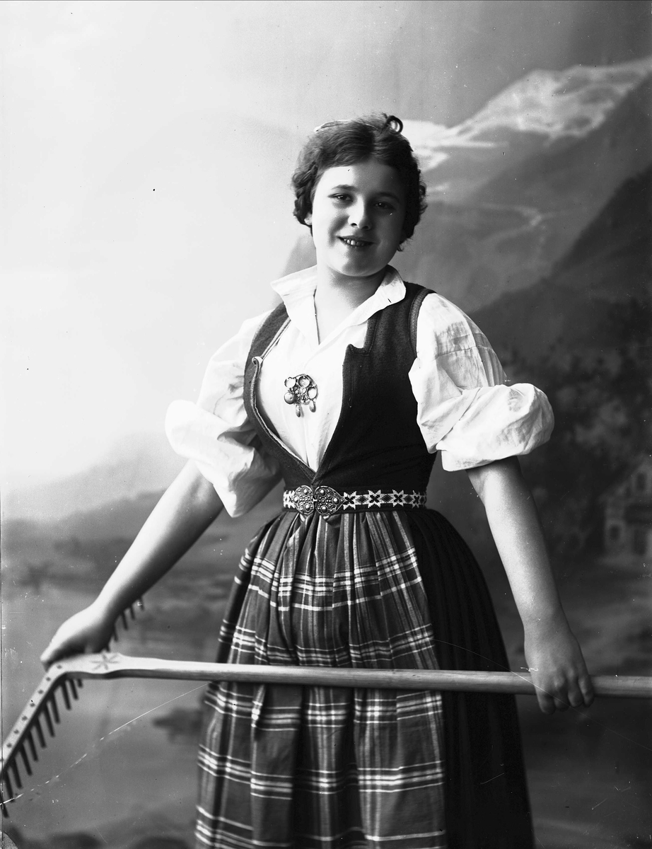 Solveig Lund, Studioportrett av kvinne i nasjonaldrakt som holder rive i hendene.Bakgrunnen er malt med fjell, fjord og hus.