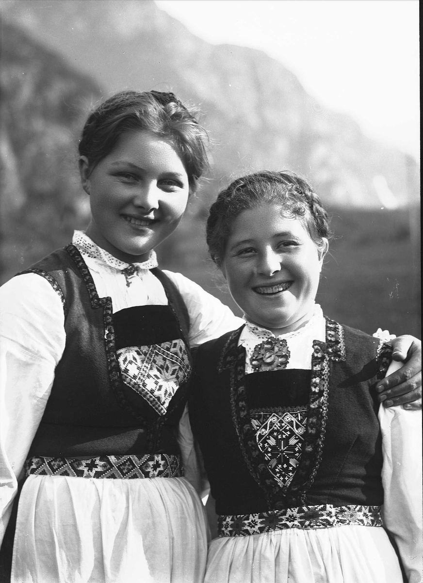 Solveig Lund, To kvinner i nasjonaldrakt holder rundt hverandre på jorde/eng. Fjell i bakgrunnen.