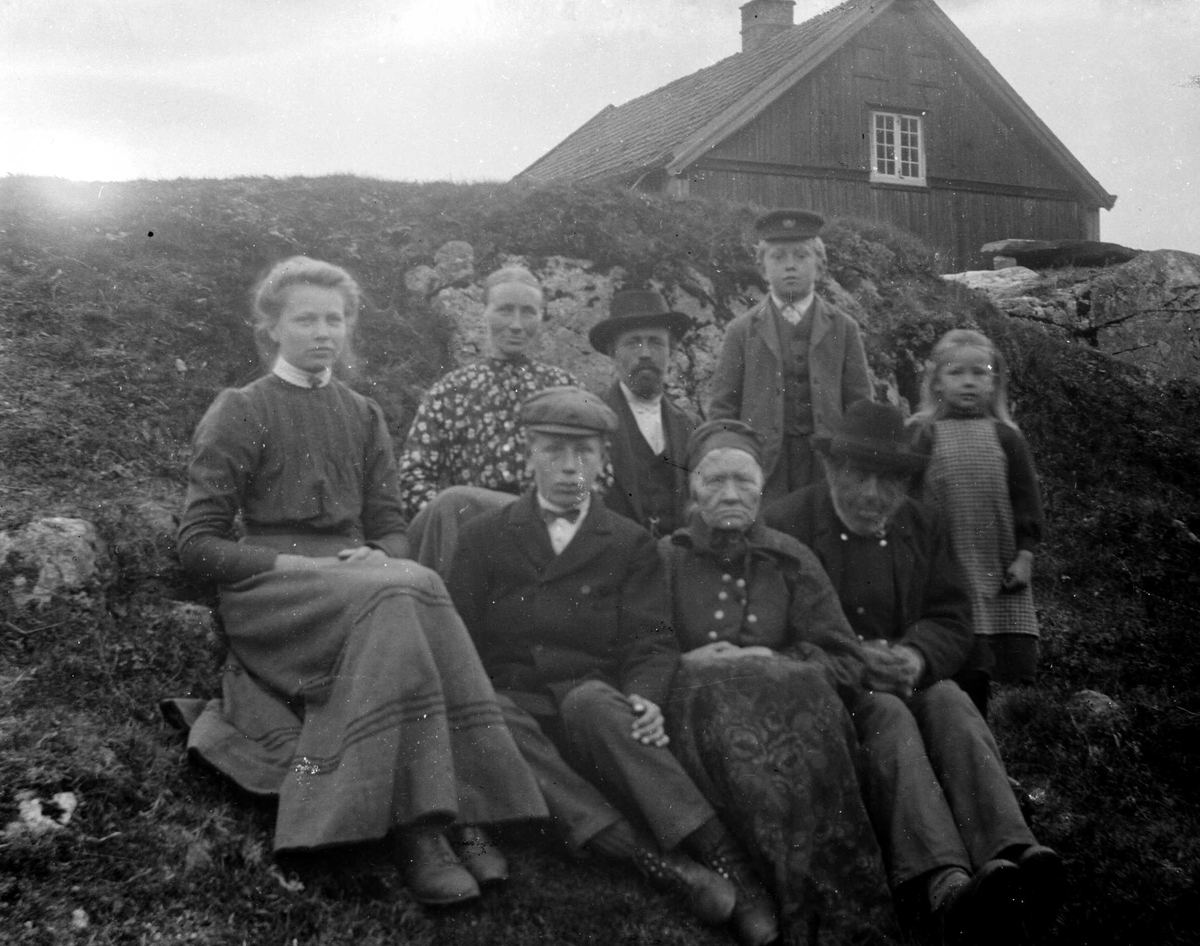 Juliane Fredrikke Langberg, Fra Eggedal, Sigdal, hos Th. Kittelsen som sitter i midten bak med hatt. Gruppe personer (8), voksne og barn, sittende i skråning foran våningshus. Eldre kvinne i folkedrakt foran. Fra malerinne Juliane Fredrikke Langbergs (1856-1930) samling, tatt ca. 1895-1910 med motivkrets slekt og venner samt …