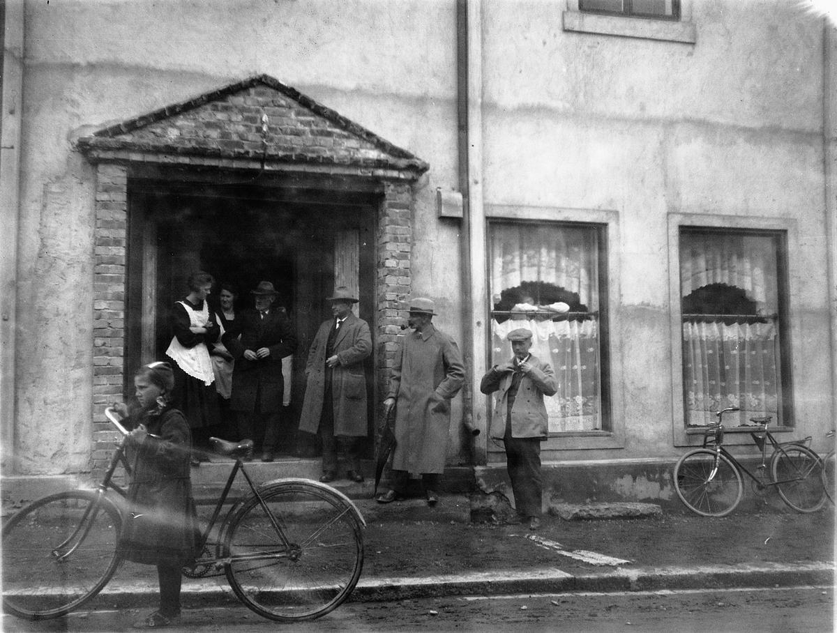 Ragnvald Andersen, Gruppe mennesker ved inngangsdøra til en kafè. Jente med en sykkel i gata.
