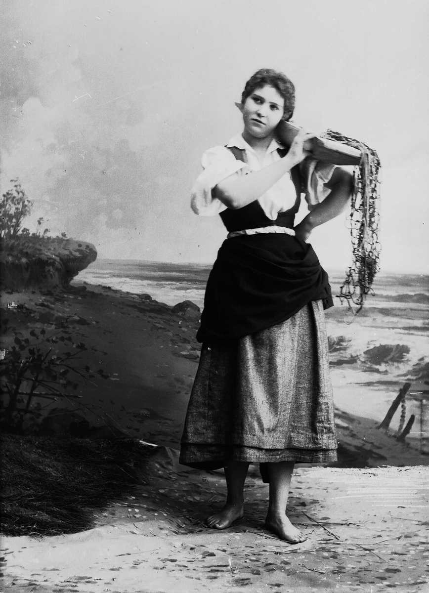 Solveig Lund, Studioportrett av kvinne i drakt som holder trau med fiskegarn på den ene skulderen. Hun ser ut mot venstre i bildet. Bakgrunnen er malt med sjø og sand.