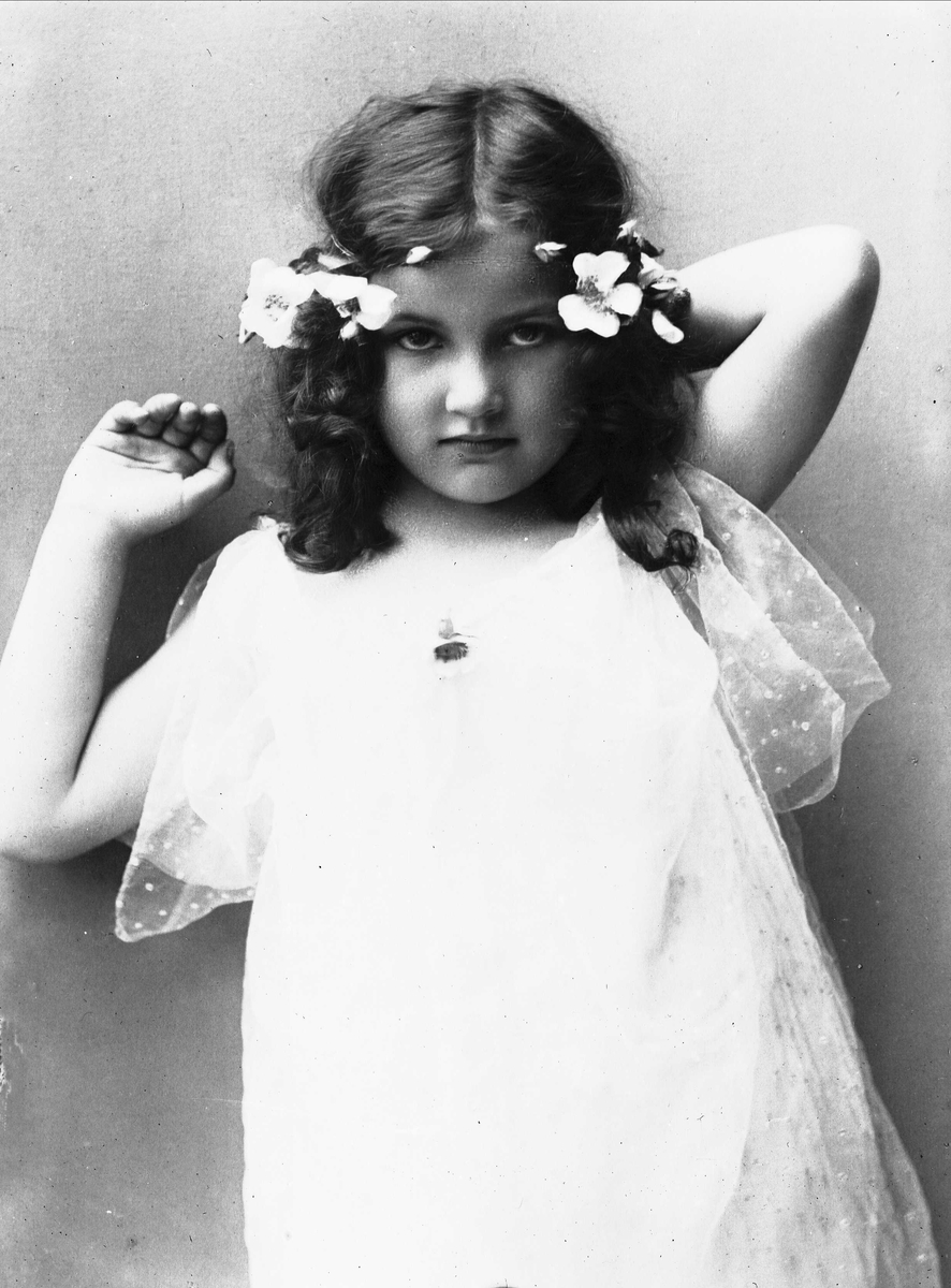Solveig Lund, 3/4 portrett av jente med kjole og blomster i håret. Løfter hendene bak hodet og ser på betrakteren.