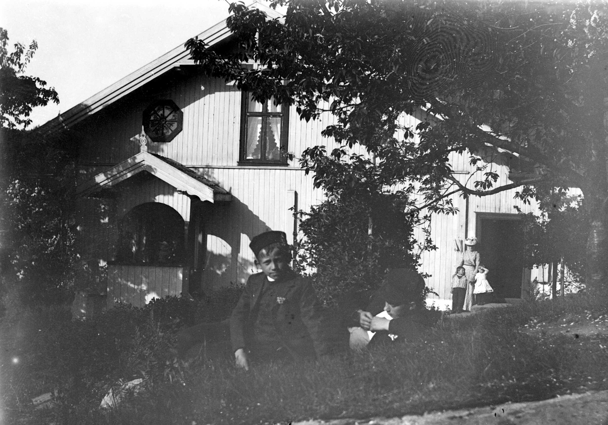 Juliane Fredrikke Langberg, Portrett av 2 gutter med lue liggende i gresset foran bolighus i sveitserstil. Guttene er sønner av klokker Busland i Hurum. der fotografens svoger var prest. Kvinne med to barn i bakgrunnen. Fra malerinne Juliane Fredrikke Langbergs (1856-1930) samling, tatt ca.1895-1910 med motivkrets slekt og ve…