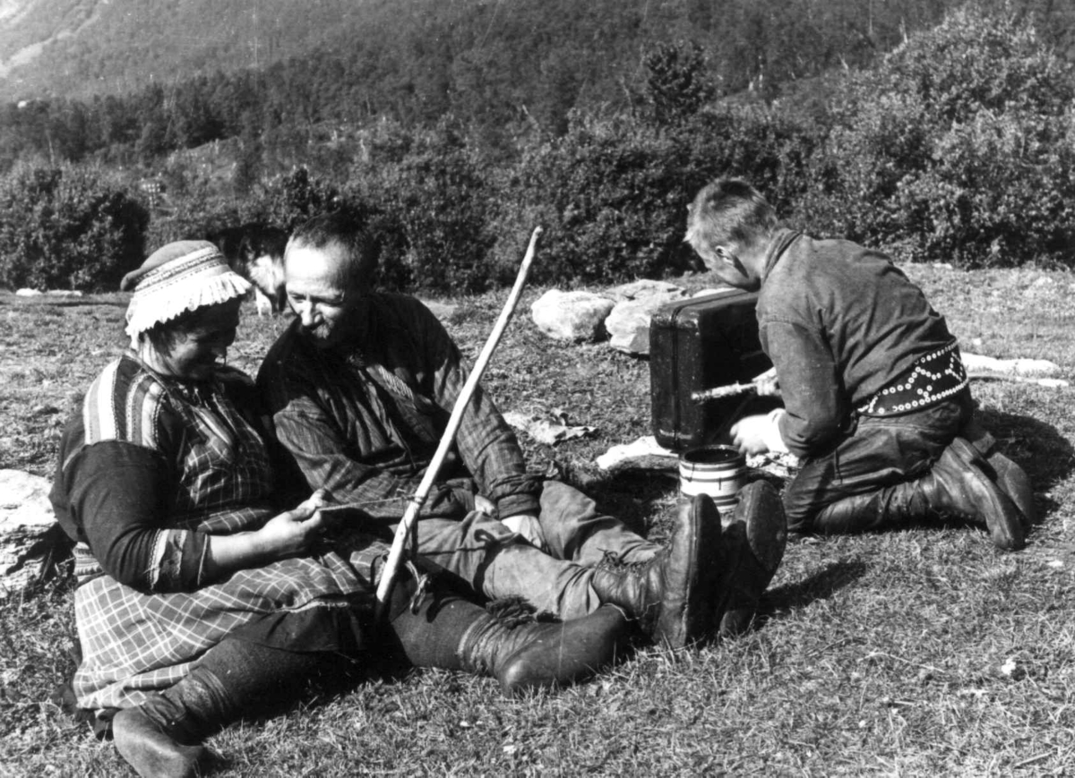 Anna Grostøl, Båndfletting. En kvinne sitter og fletter bånd, to menn ved siden av henne. Lyngen, Troms 1947.