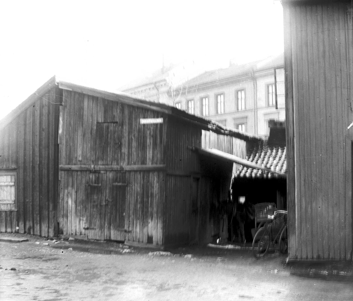 Juliane Fredrikke Langberg, Fra uthus (garasje? )i bakgård (?) med bygård i bakgrunnen,ukjent sted. Parkert sykkel. Noe uskarpt bilde. Fra malerinne Juliane Fredrikke Langbergs (1856-1930) samling, tatt ca.1895-1910 med motivkrets slekt og venner samt steder tilknyttet samvær med disse.