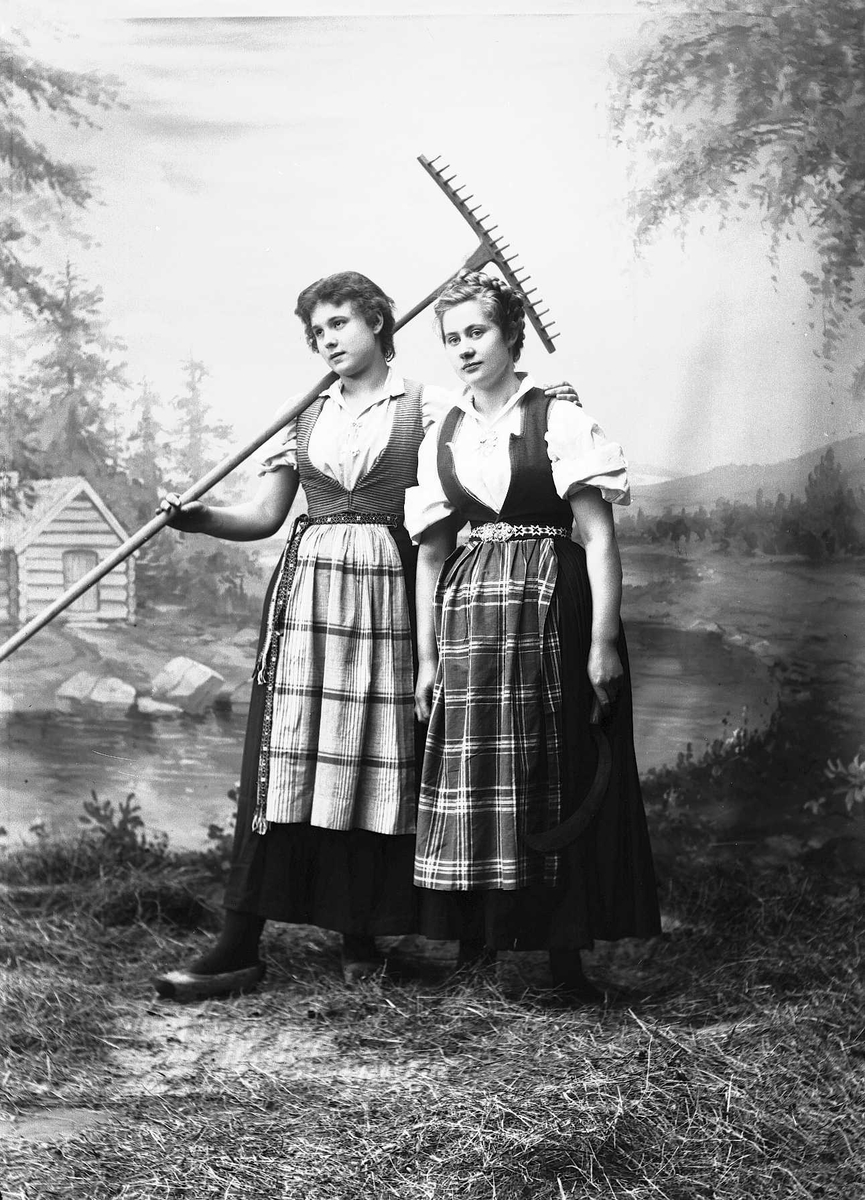 Solveig Lund, Studioportrett av to kvinner i nasjonaldrakter som holder hverandre i hendene.Den ene holder en rive over skulderen. Bakgrunnen er malt med skog, vann og hytte. På gulvet er det høy/gress.