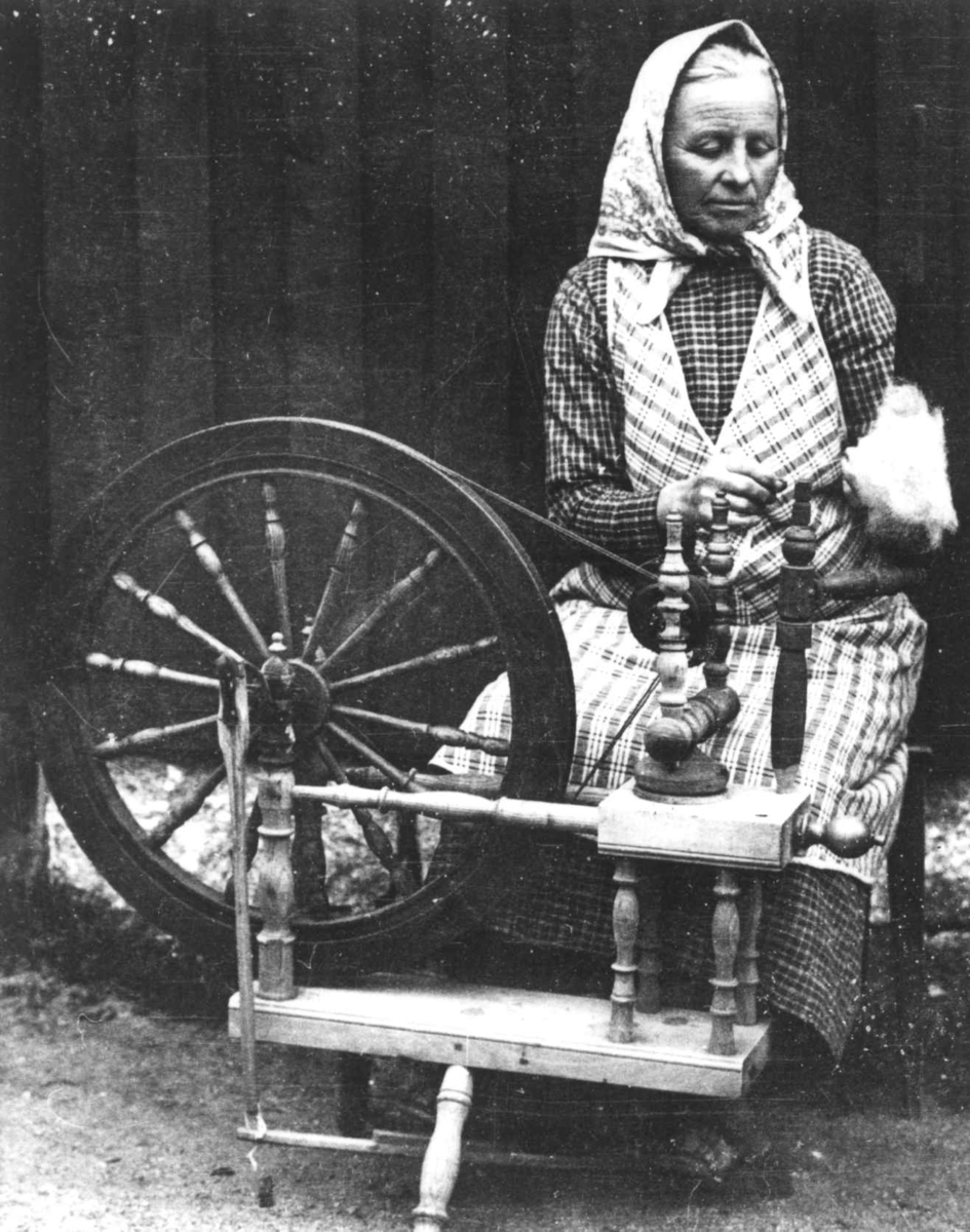 Anna Grostøl, En kvinne i rutete kjole, forkle og skaut sitter ved en rokk og spinner. Ukjent sted 1936.